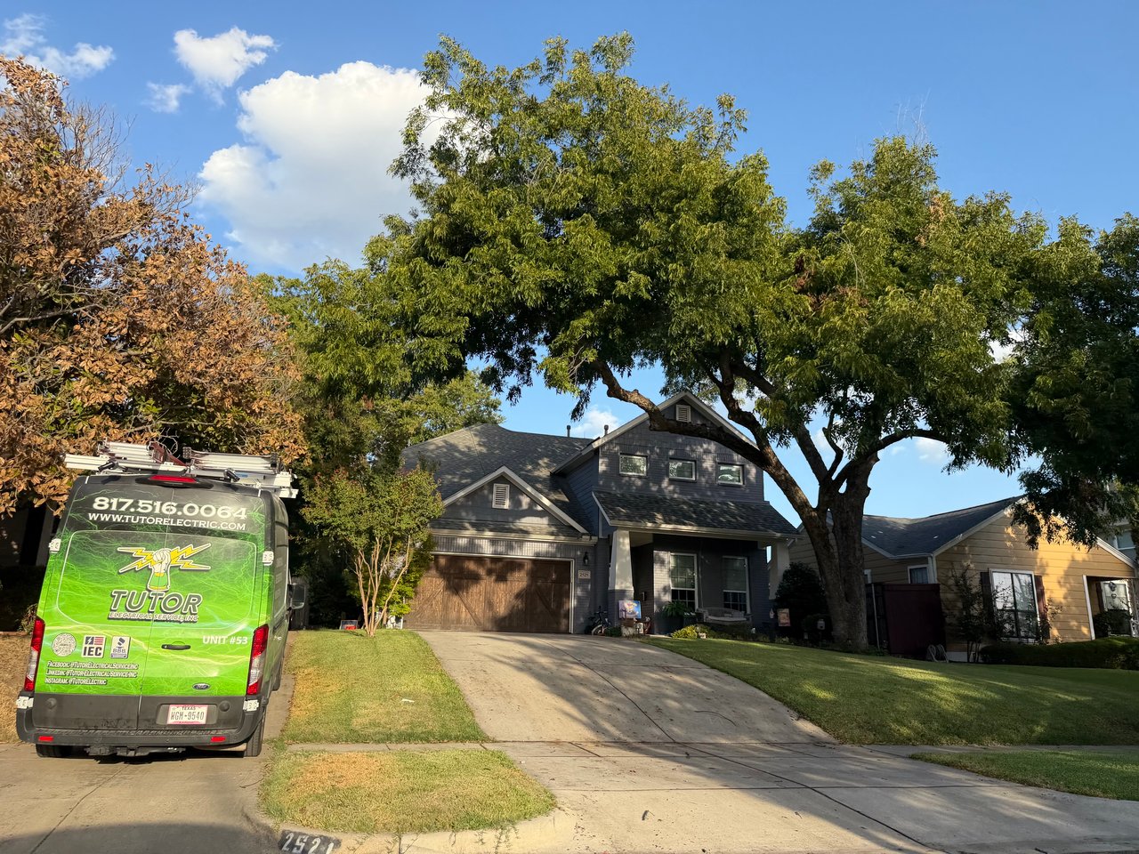 Two-story gray house beneath mature trees, with a driveway and green service van.