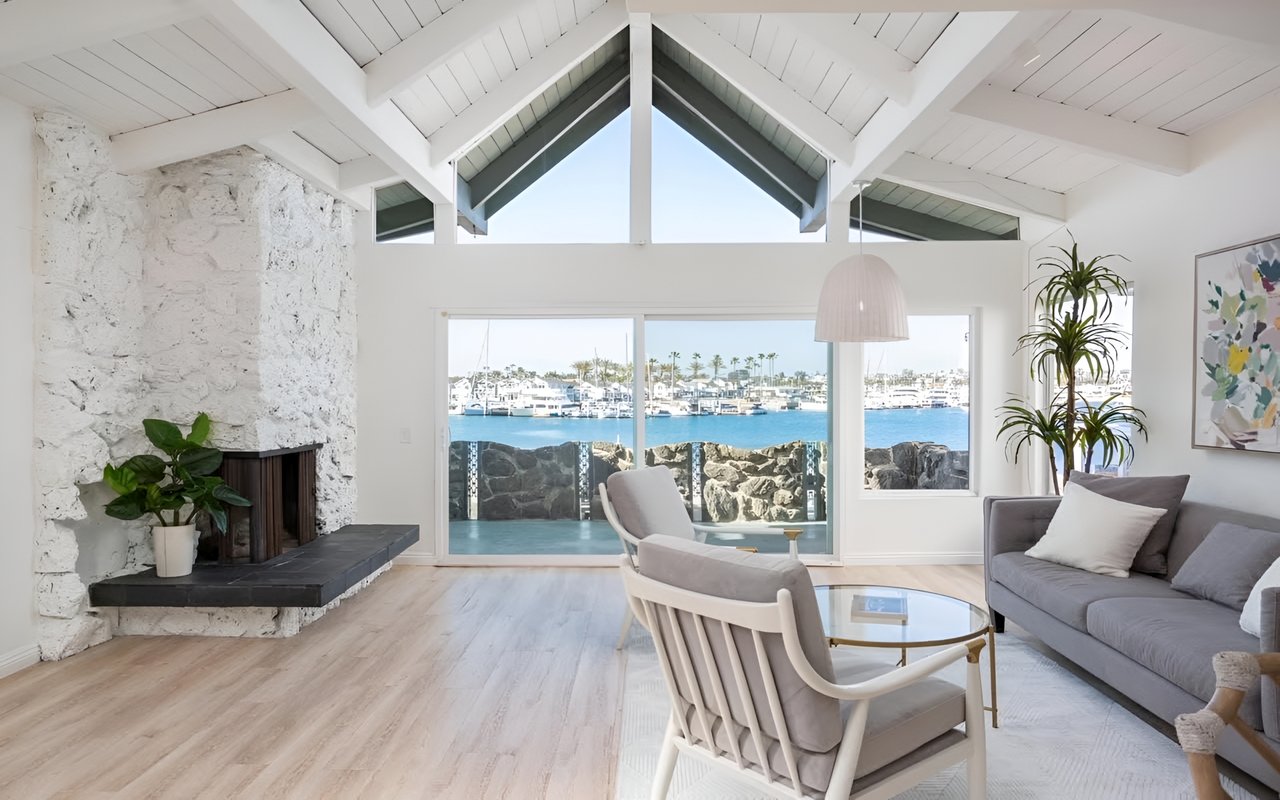  A bright living room with white vaulted ceilings and a stone fireplace. Large windows reveal a marina filled with boats.