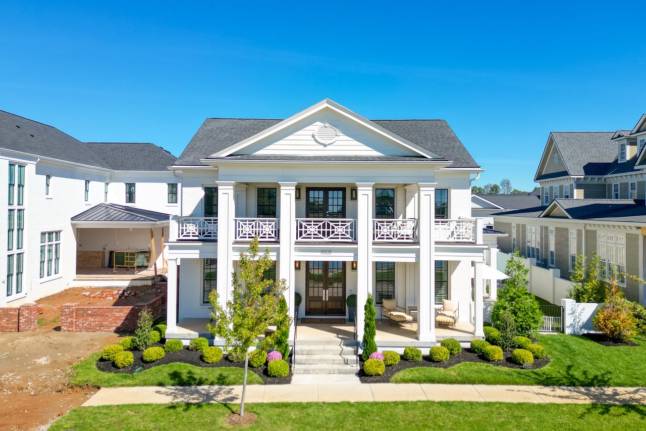 White two-story house with columned porch, upper balconies, and landscaped front lawn.