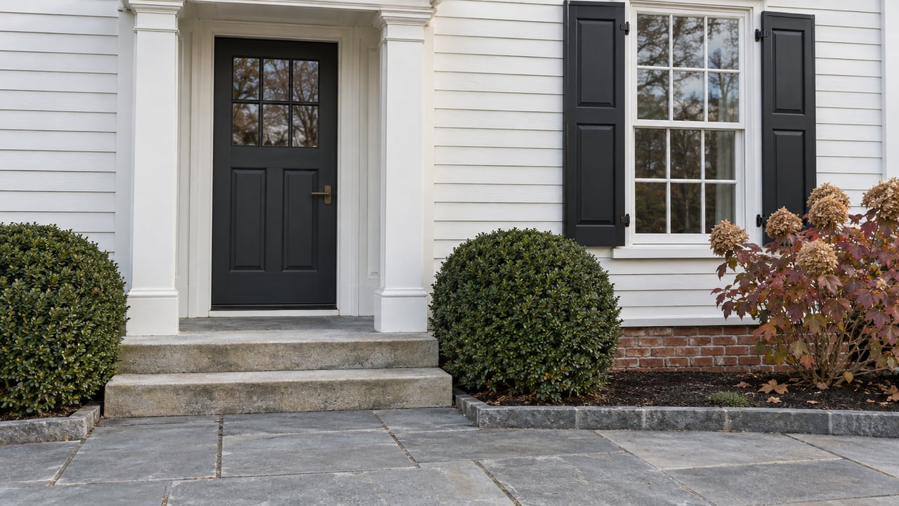 White clapboard entrance with a charcoal door, paired black shutters, worn stone steps and clipped boxwood beside autumn oakleaf hydrangea.
