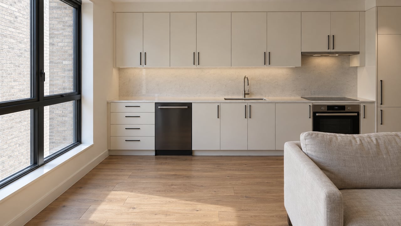 Compact condominium kitchen with cream cabinets, black-framed window facing brick, and oak flooring beside an oatmeal sofa.