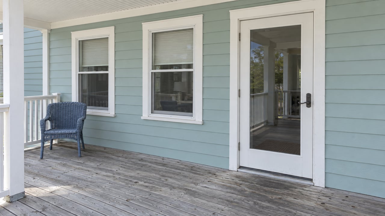 Seafoam raised-house entry with white trim, weathered deck boards and a faded blue outdoor chair beneath a covered porch.
