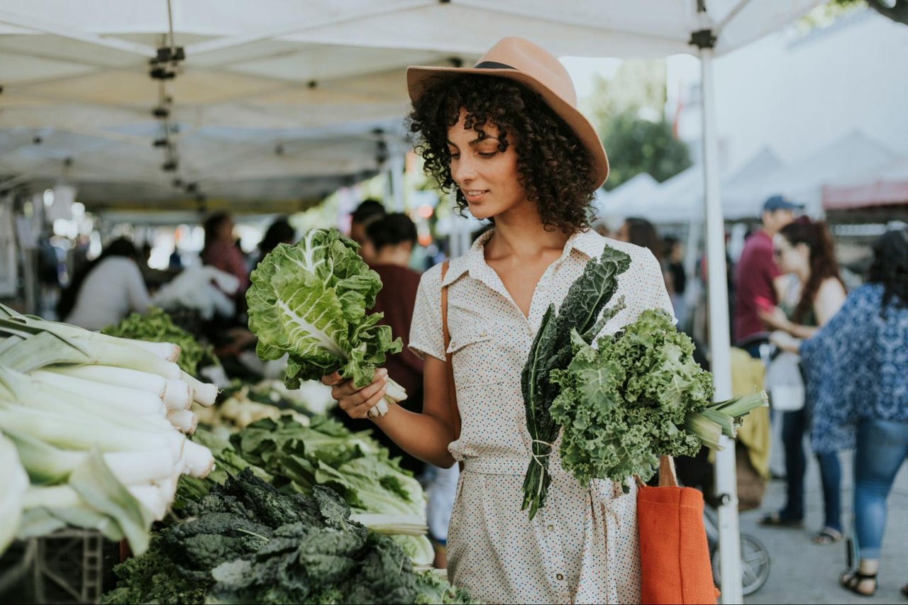 Woman in a wide-brimmed hat shopping for leafy greens at an outdoor farmers market.