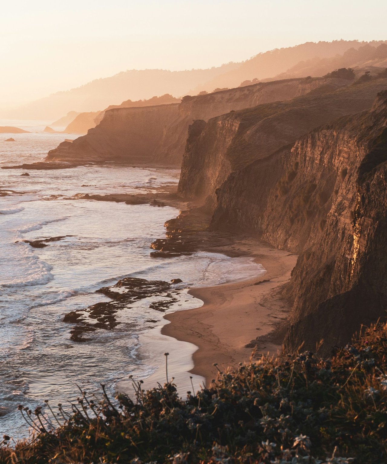 Waves meet a narrow beach beneath rugged cliffs along the California coast at golden hour.