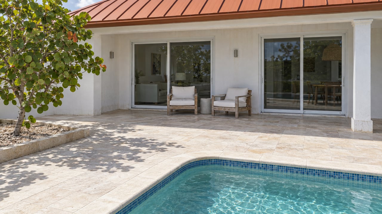 Sunlit white stucco home with a red-orange metal roof, limestone pool terrace, teak chairs, and a sculptural sea grape tree.