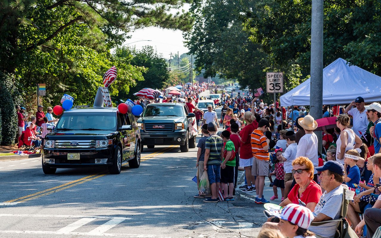 A community parade passes spectators on a shaded, tree-lined street.