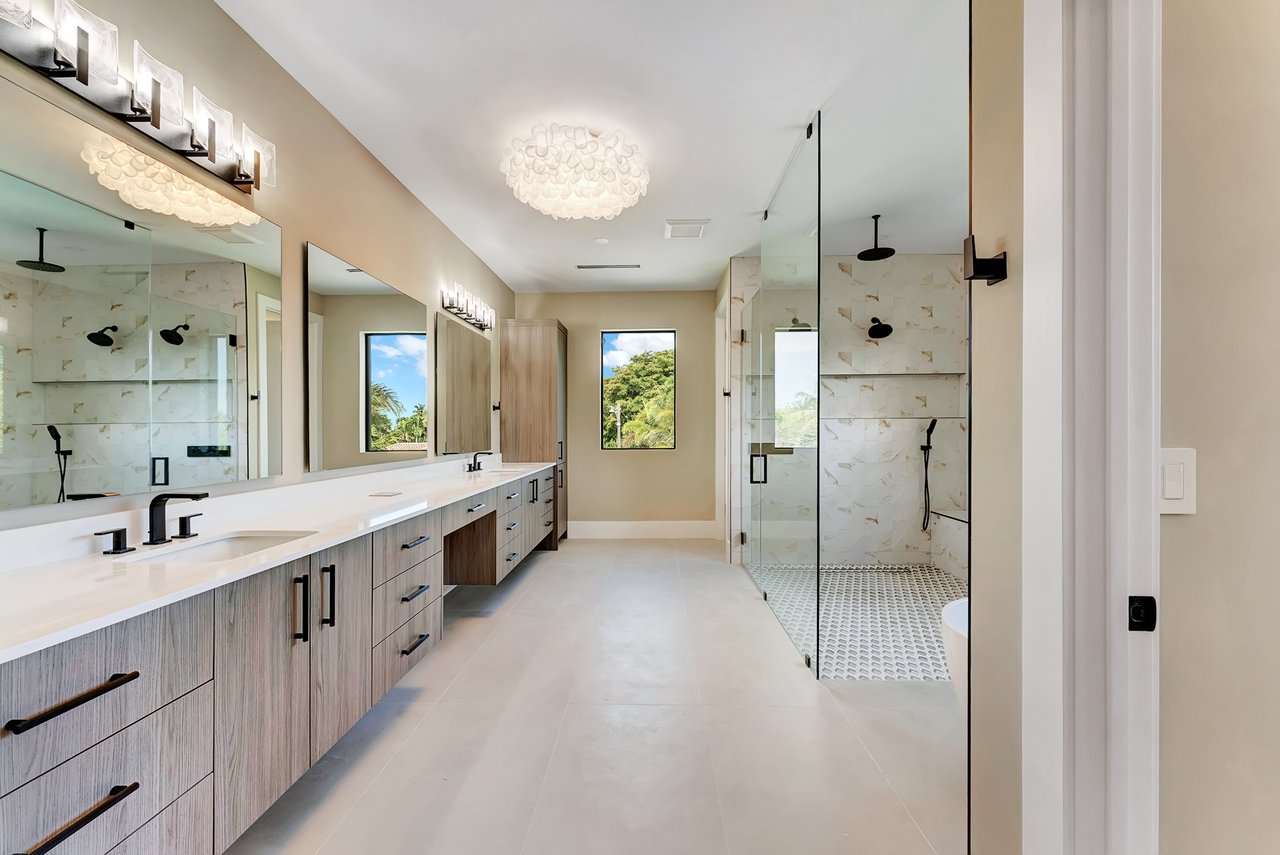 Bathroom with long double vanity, glass-enclosed shower, and two windows overlooking greenery.