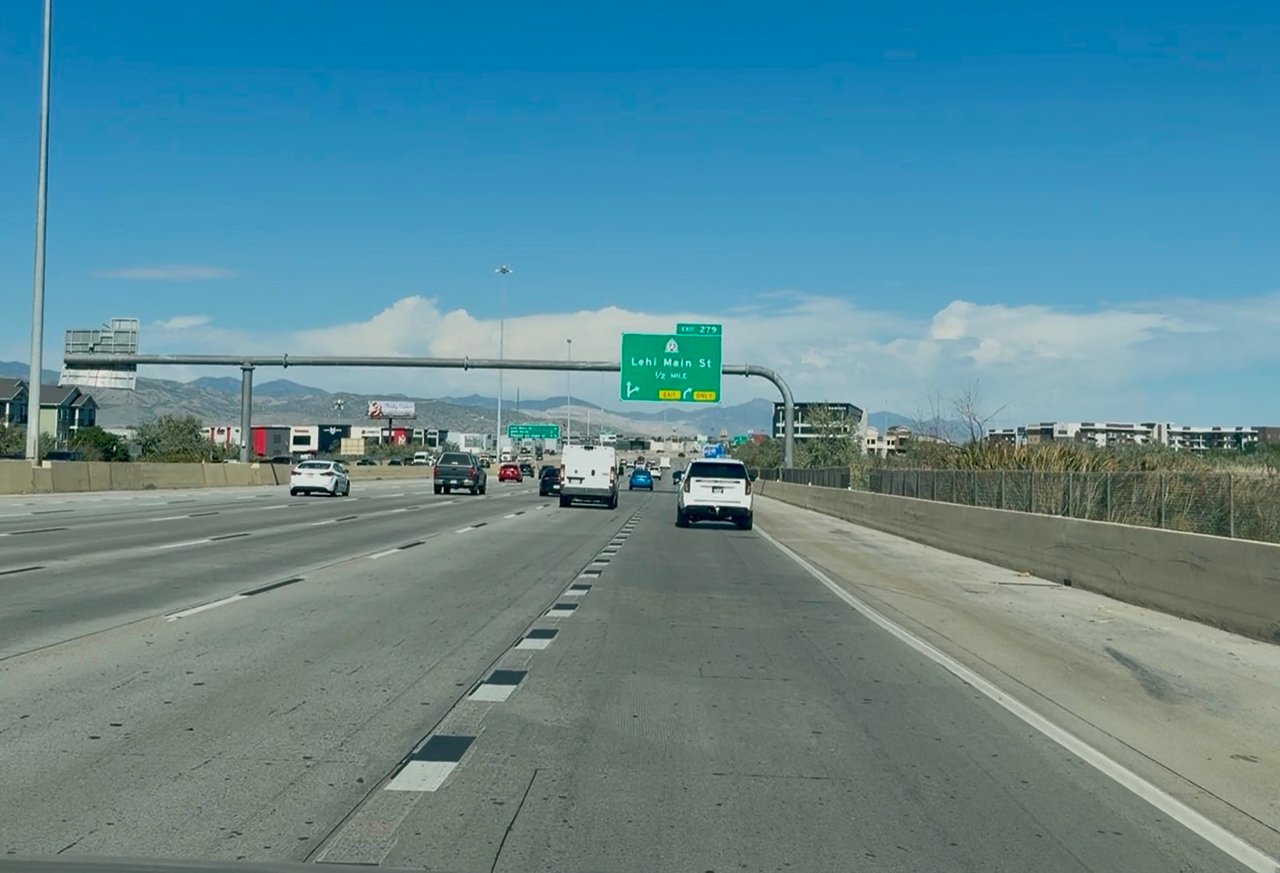 Highway traffic approaches the Lehi Main Street exit beneath a blue sky.