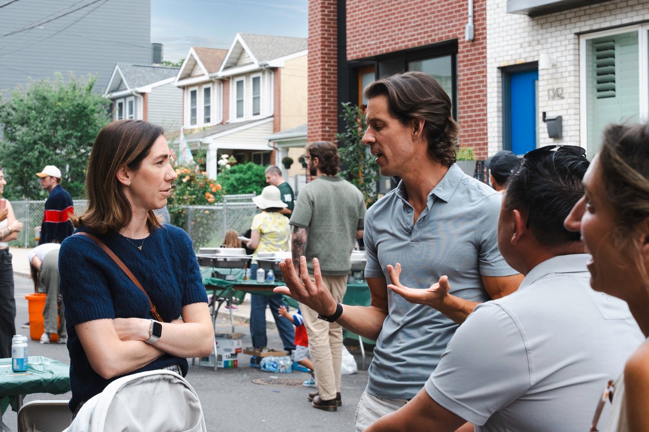 Neighbors chat at a block party on a residential street beside folding tables and refreshments.