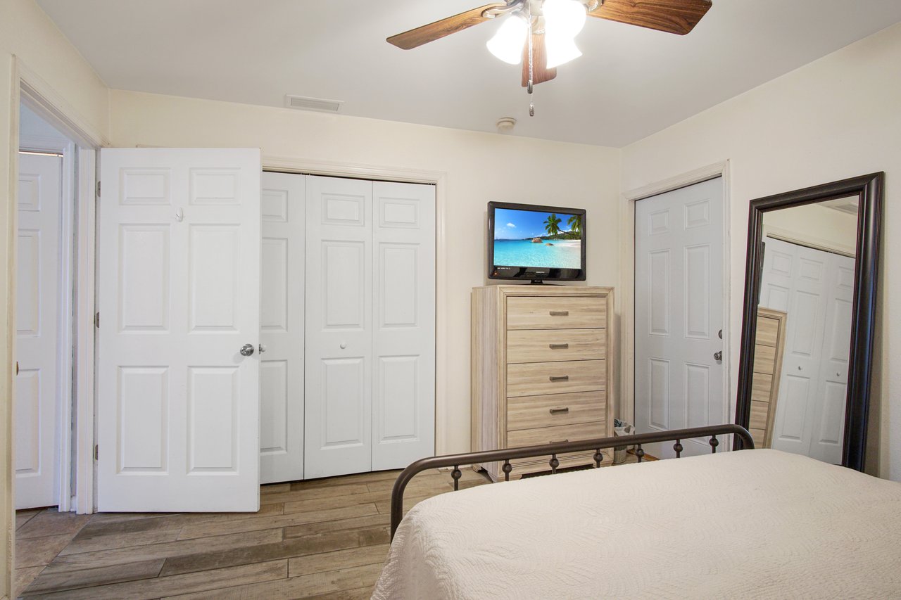 Bedroom with light walls, ceiling fan, and wood floor. Features include a TV on a wooden dresser, a large mirror, open closet, and metal bed frame. Cozy and simple.
