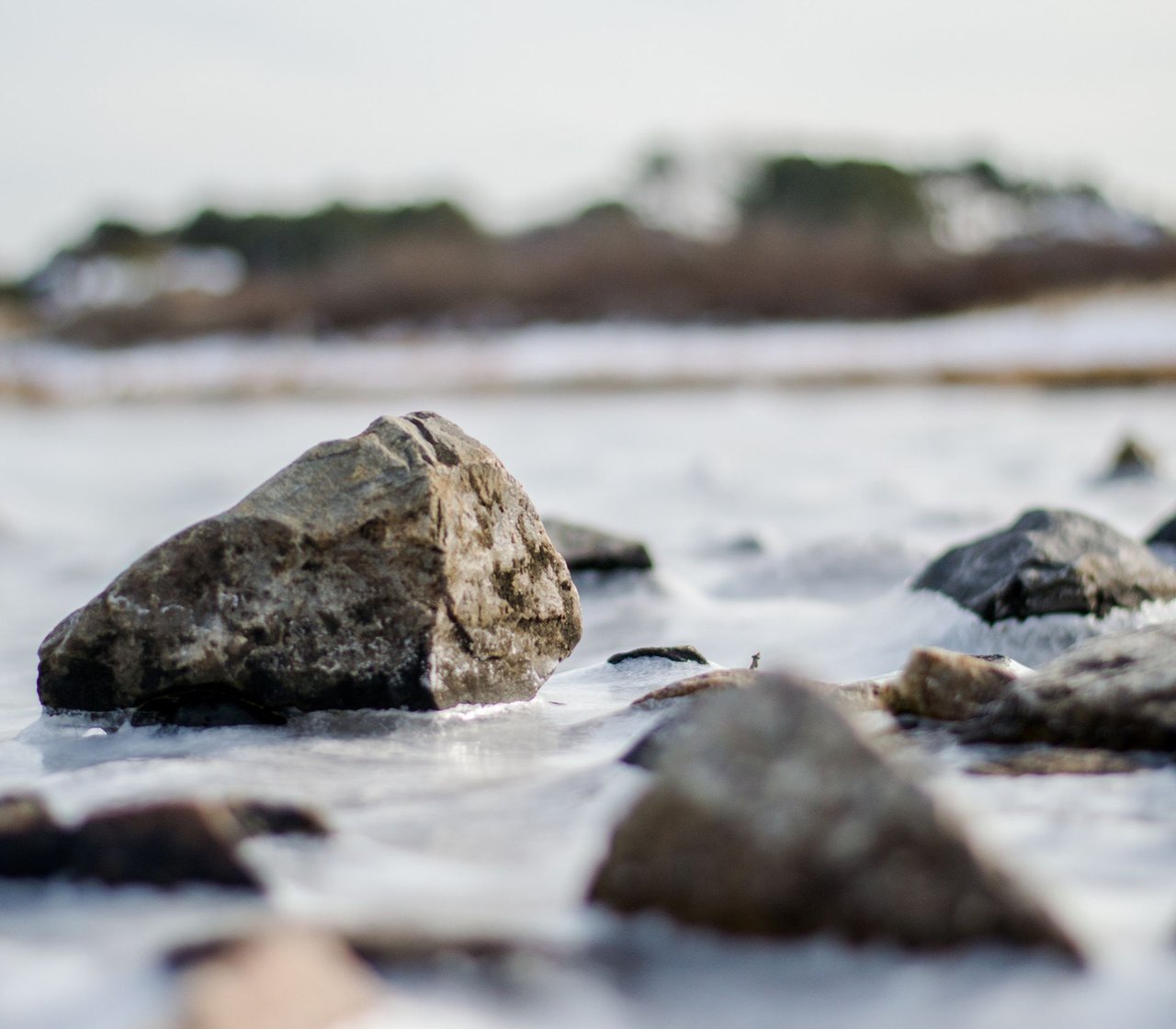 Large rocks sit in shallow surf along a shoreline, with the distant coast out of focus.