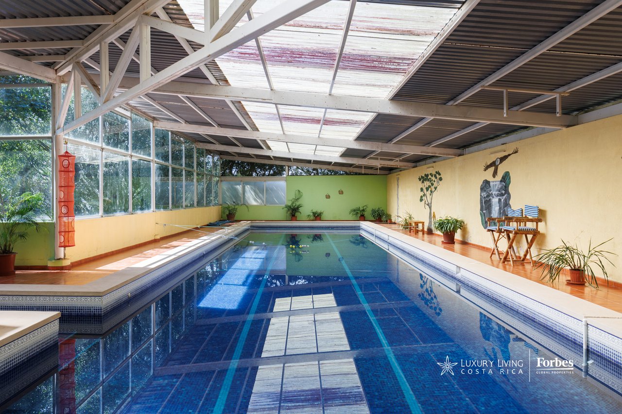 Indoor swimming pool beneath a high roof, with glass walls and tiled decking.