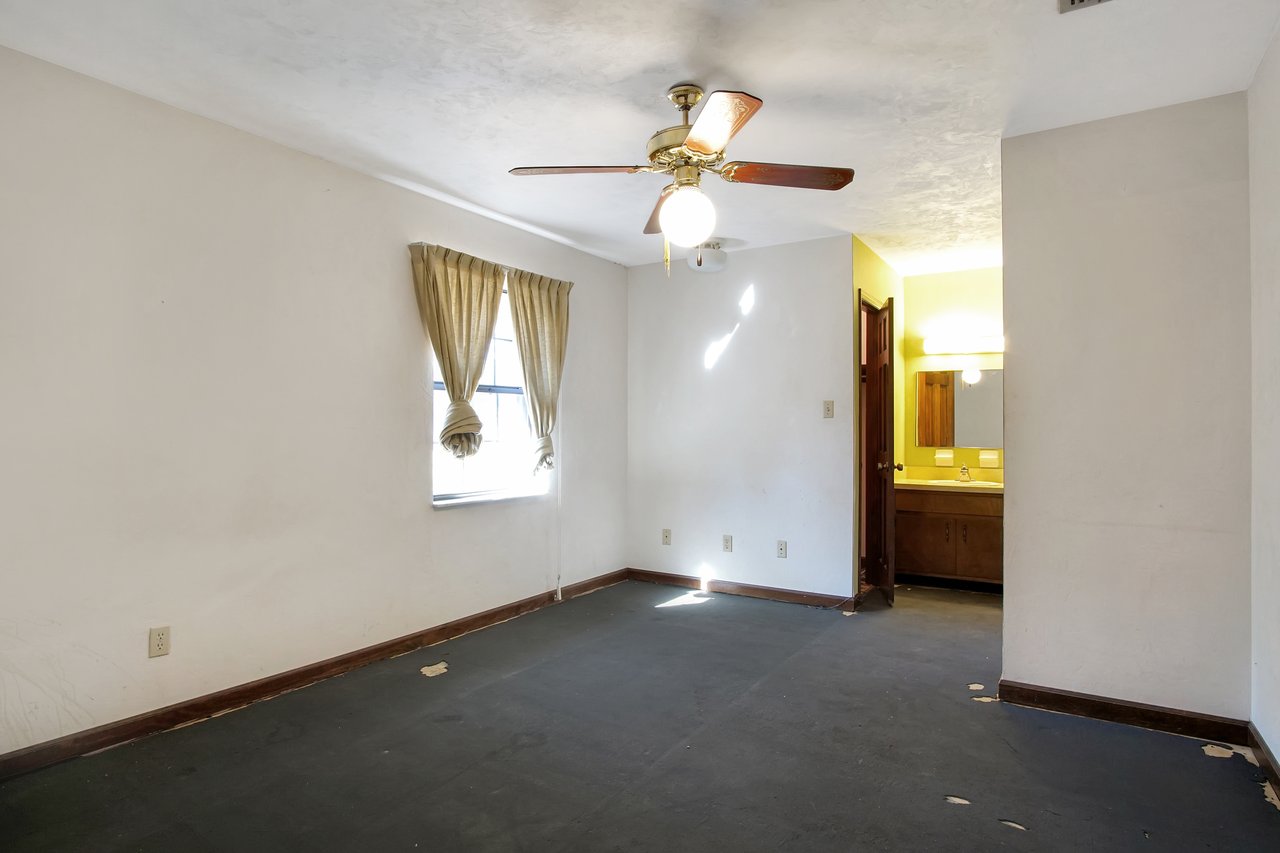 Empty, dim-lit room with worn carpet and a ceiling fan. A window with beige curtains lets in light. An open door reveals a bathroom with a lit mirror.