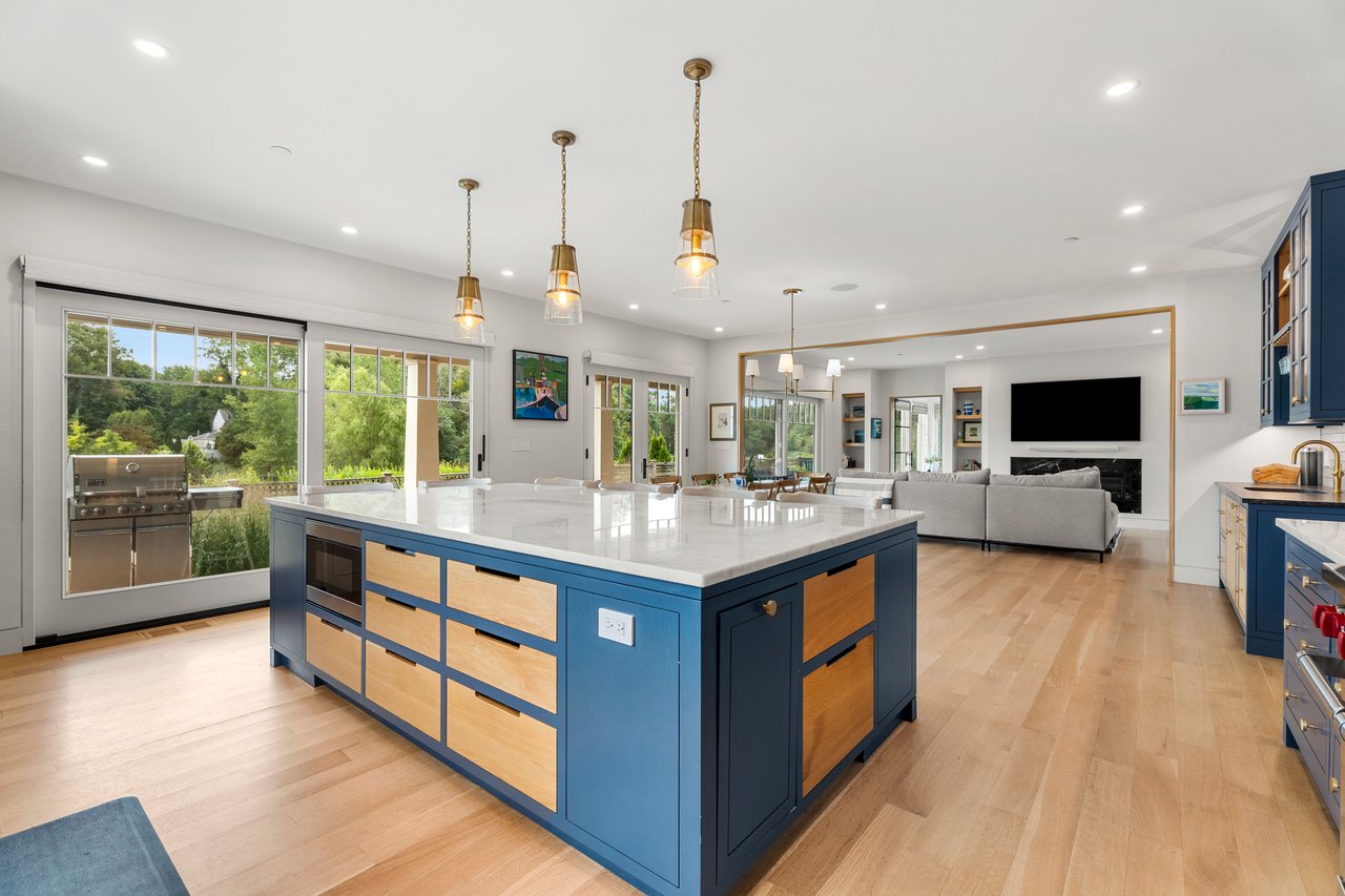 Open-plan kitchen with a blue-and-wood island, brass pendants, and large windows overlooking greenery.
