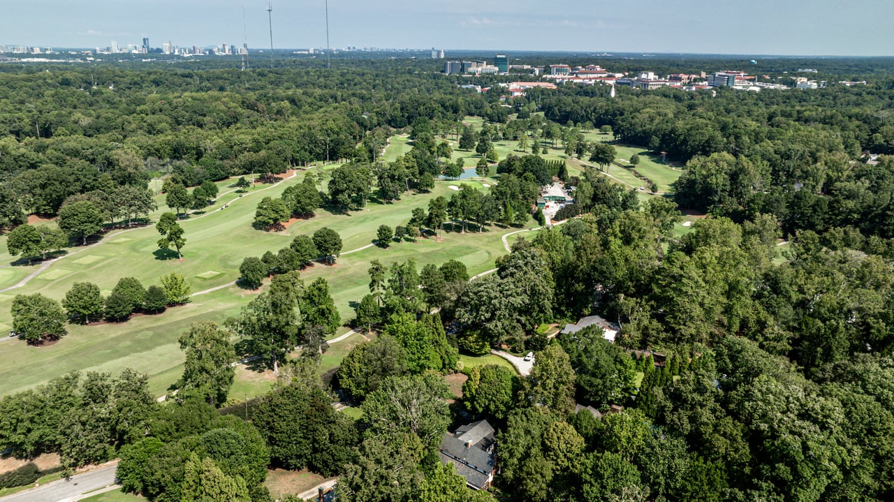 Aerial golf course bordered by wooded homes, with a distant city skyline.