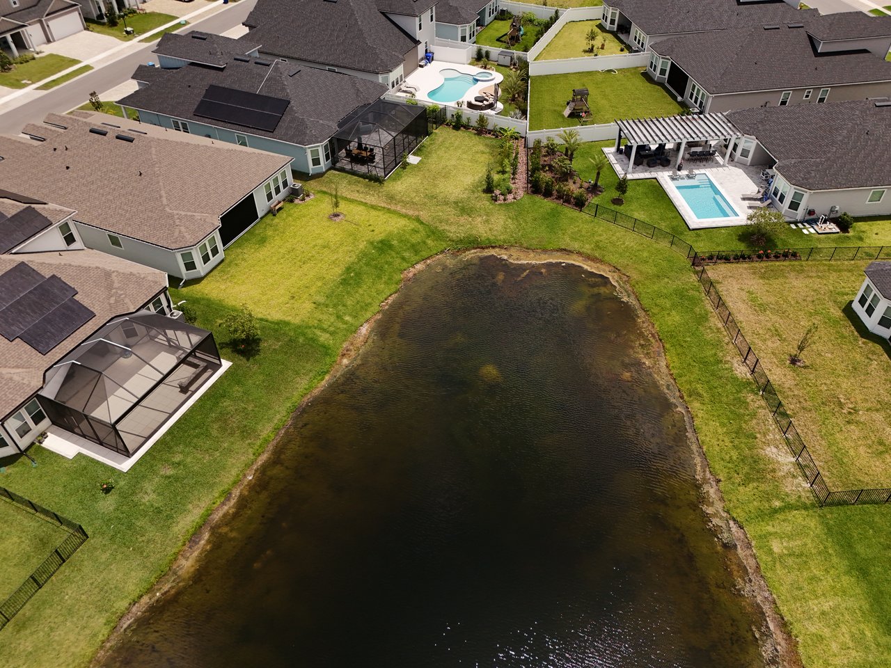 Aerial view of homes around a pond, with fenced yards, screened patios, and swimming pools.