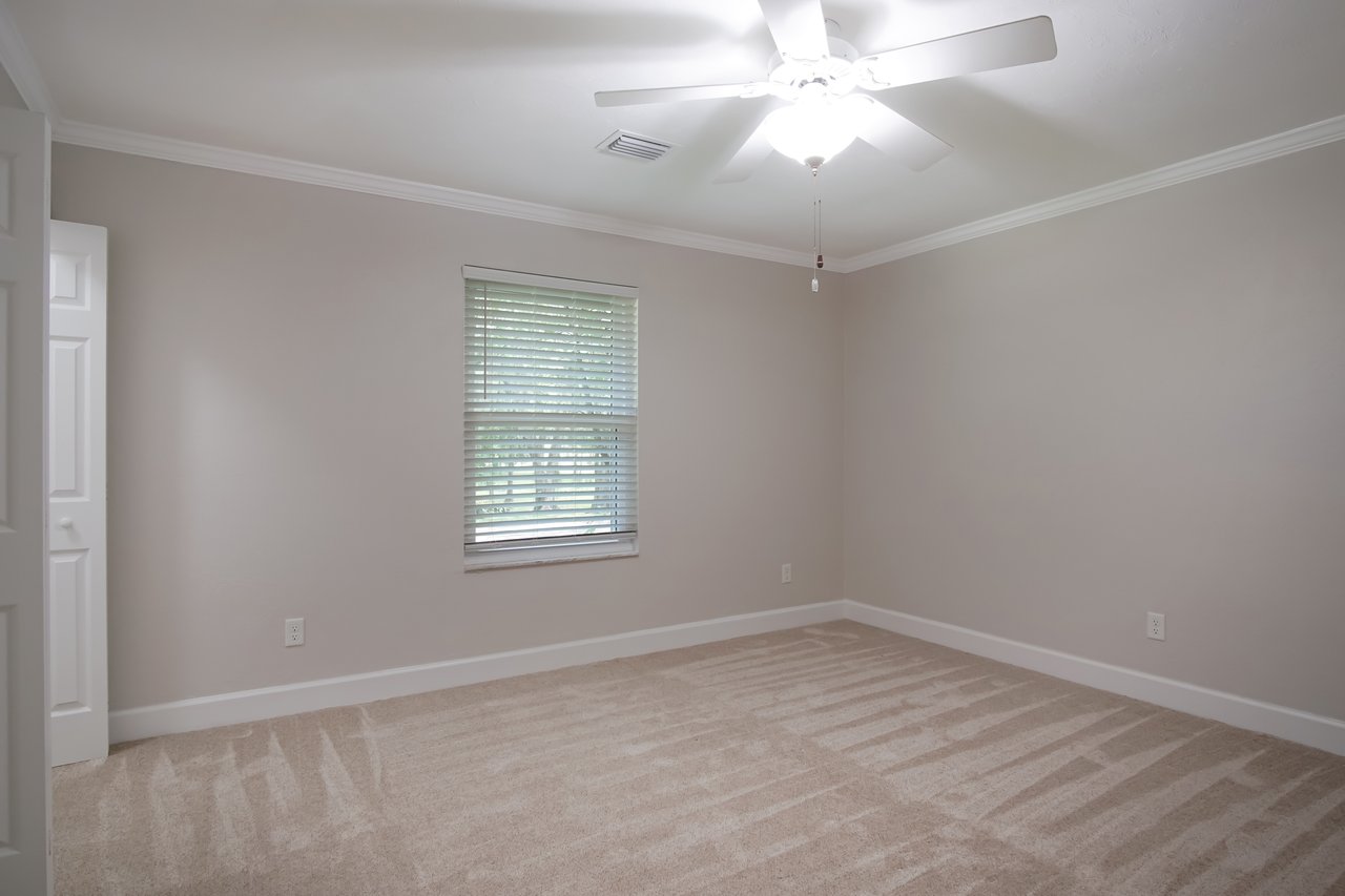 Empty room with beige walls and carpet. A window with blinds lets in soft light, and a white ceiling fan with light fixture hangs overhead. Calm, minimalist feel.