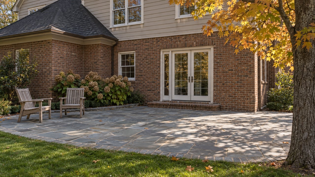 Autumn sunlight on a brick-and-taupe-sided home's stone terrace, with two teak chairs and a golden sugar maple.