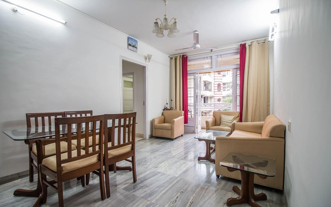 Furnished living and dining room with beige sofas, wooden dining chairs, and marble flooring.