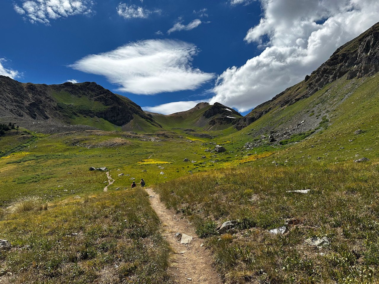 A trail winds through a green alpine meadow toward a rocky mountain valley beneath dramatic clouds.