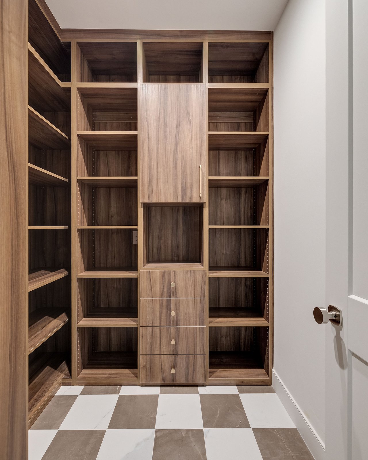 Walk-in closet with walnut built-in shelving, central drawers, and cream-and-taupe checkerboard tile flooring.
