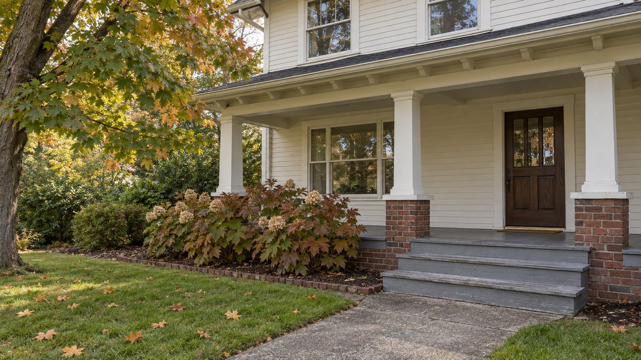 An ivory wood-sided house with a covered timber porch, brick foundation, autumn maple canopy and broad lawn.