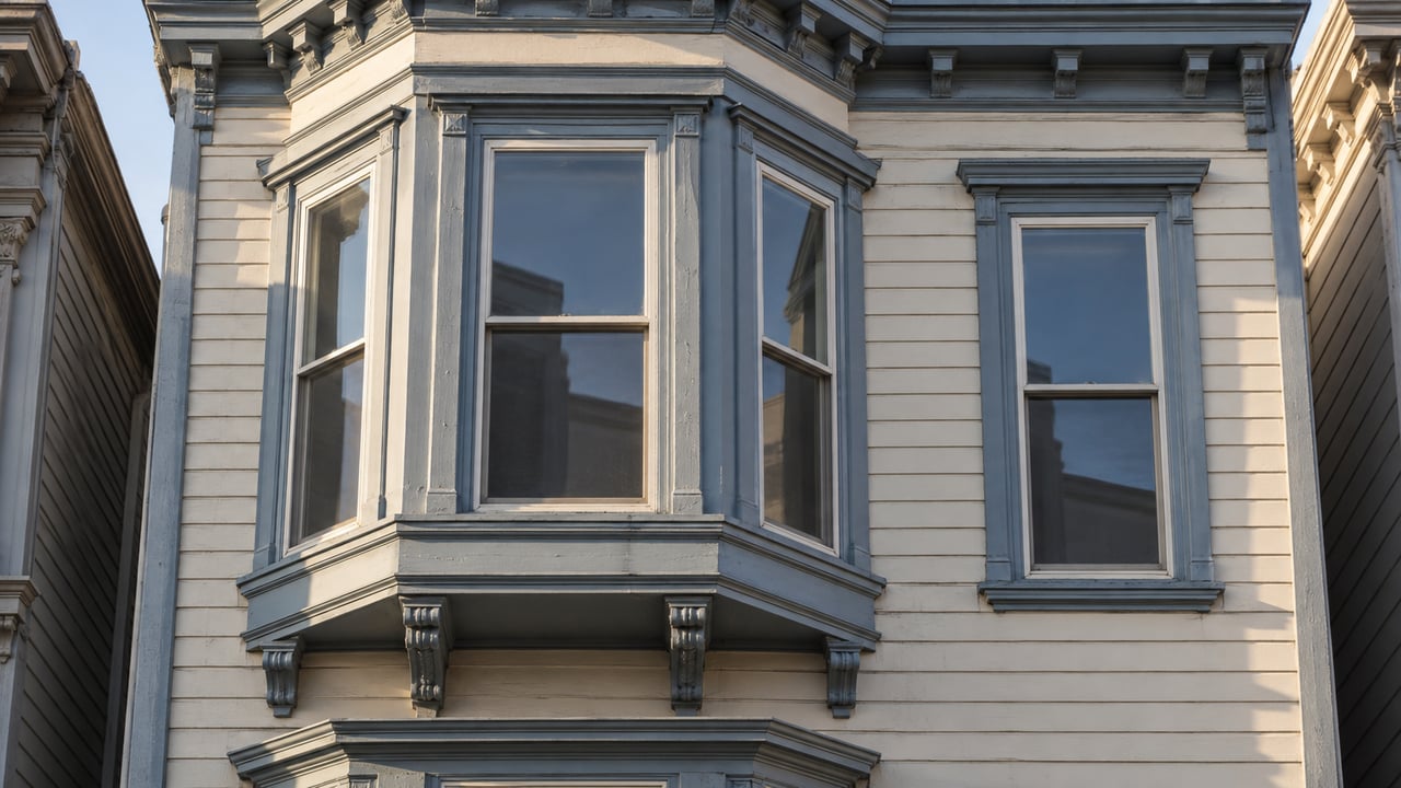 Close view of a cream-painted wood facade with blue-gray bay-window trim, carved brackets, and gently weathered siding in afternoon sunlight.