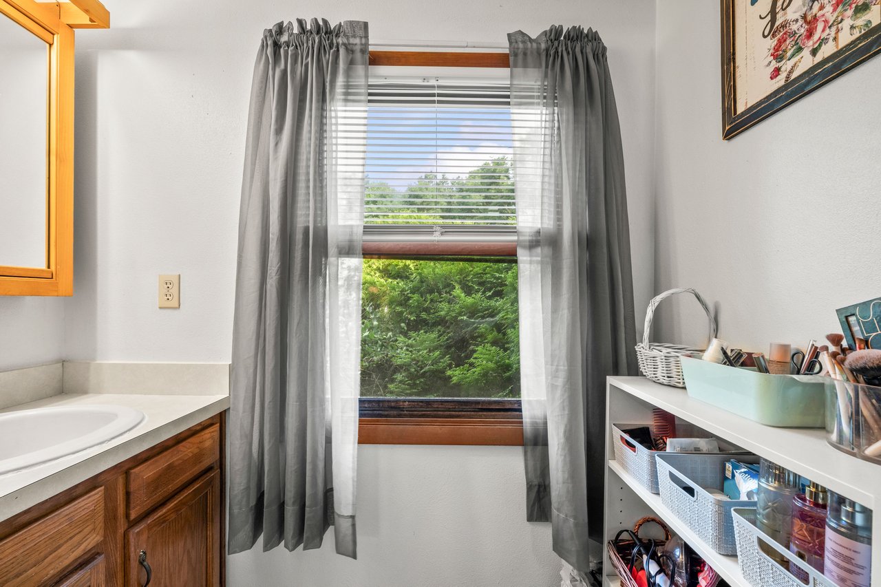 Bathroom vanity beside a curtained window and open shelving stocked with toiletries.