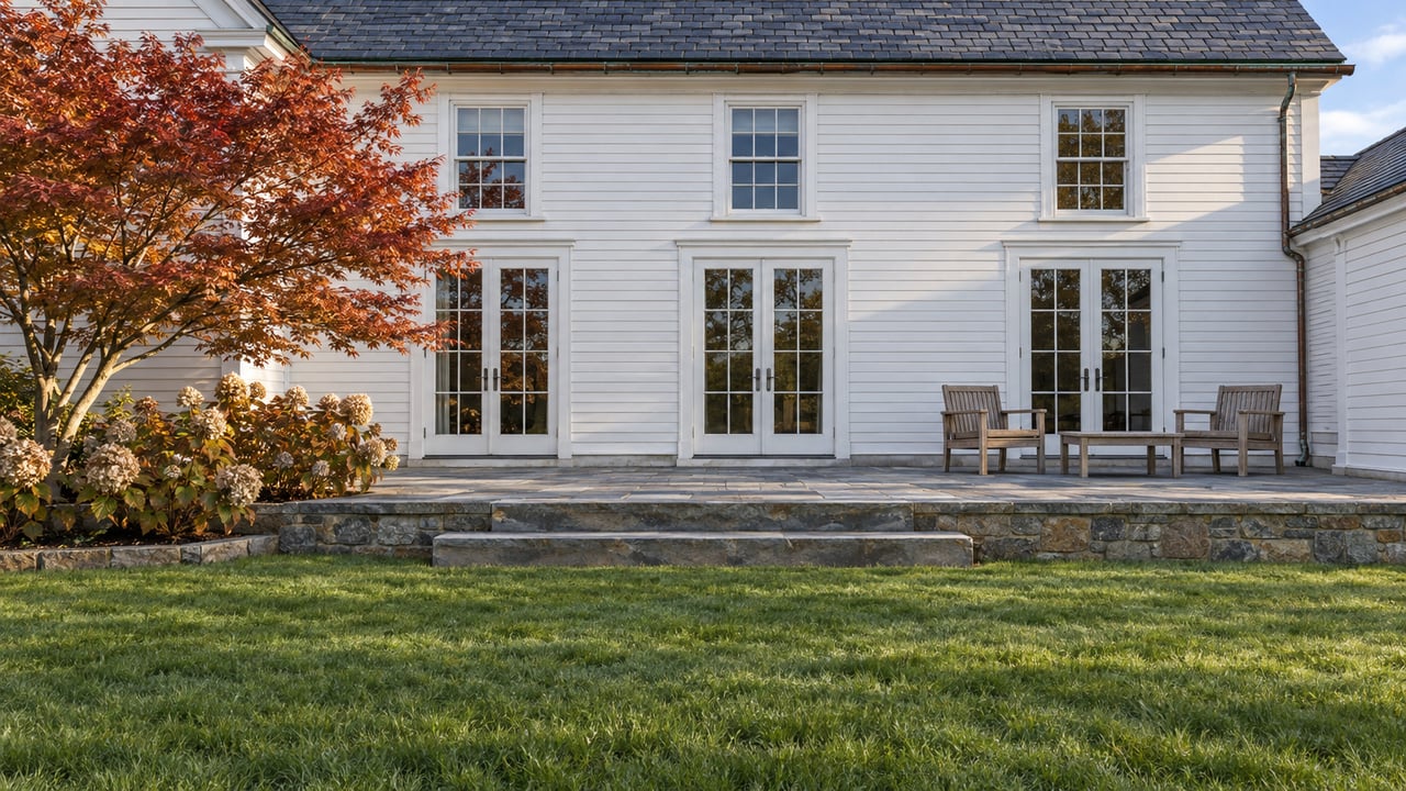 White clapboard home with tall glazed doors, slate roof and a sandstone terrace overlooking a deep green lawn, with a Japanese maple and oakleaf hydrangeas.