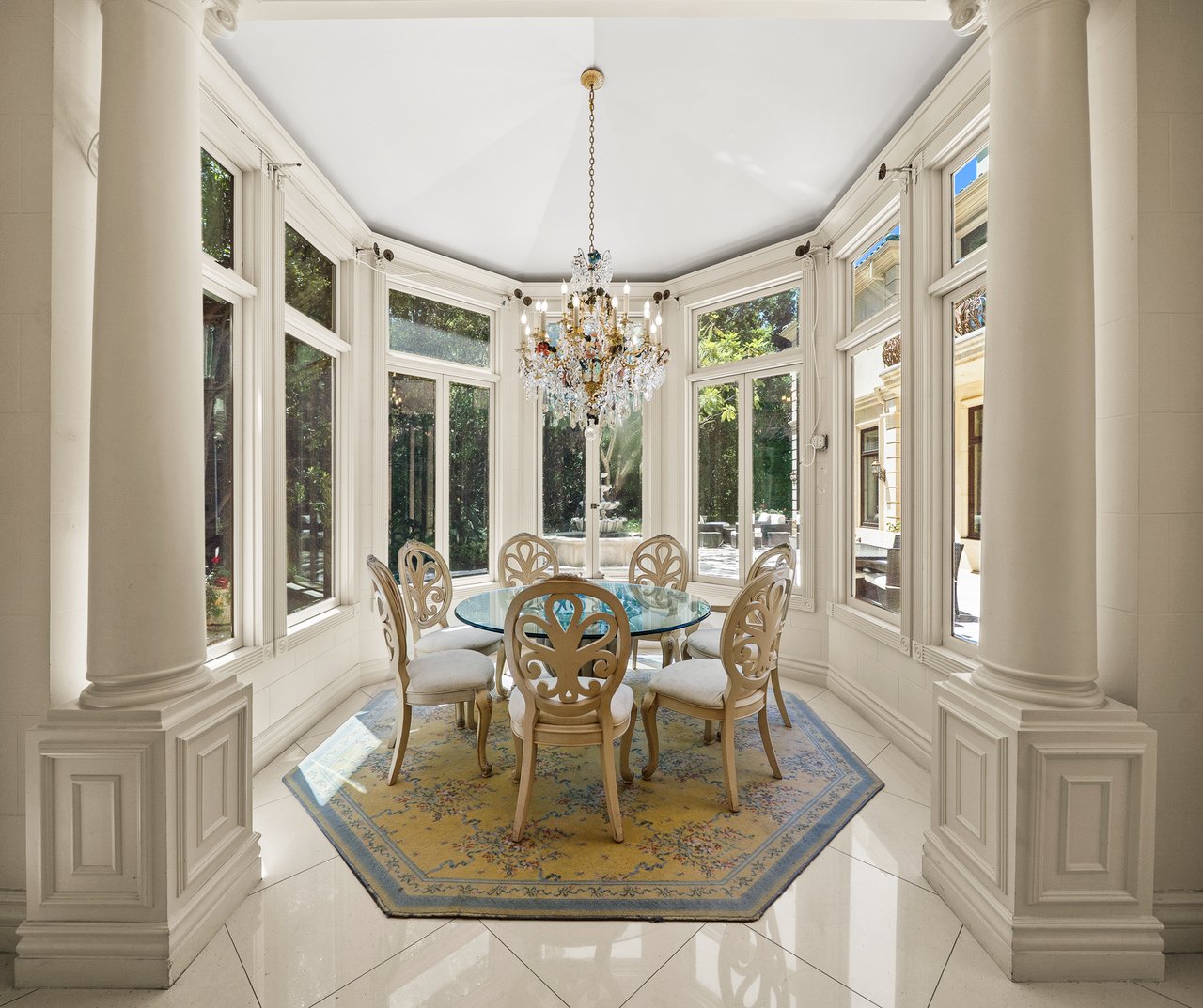 Formal dining room with a glass-top table, crystal chandelier, tall windows, and white columns.