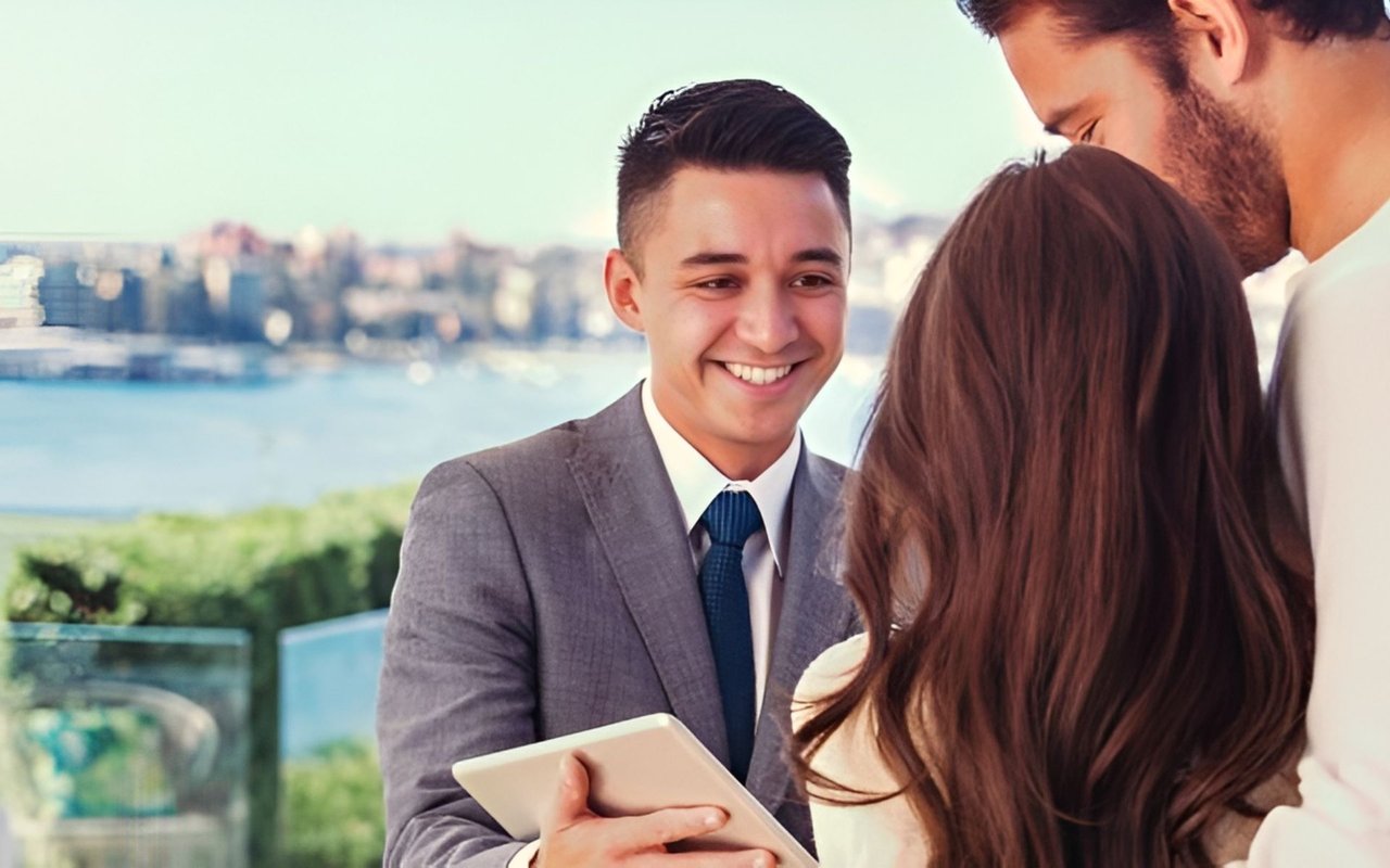 A suited man holding a tablet speaks with a woman and another man beside a waterfront.