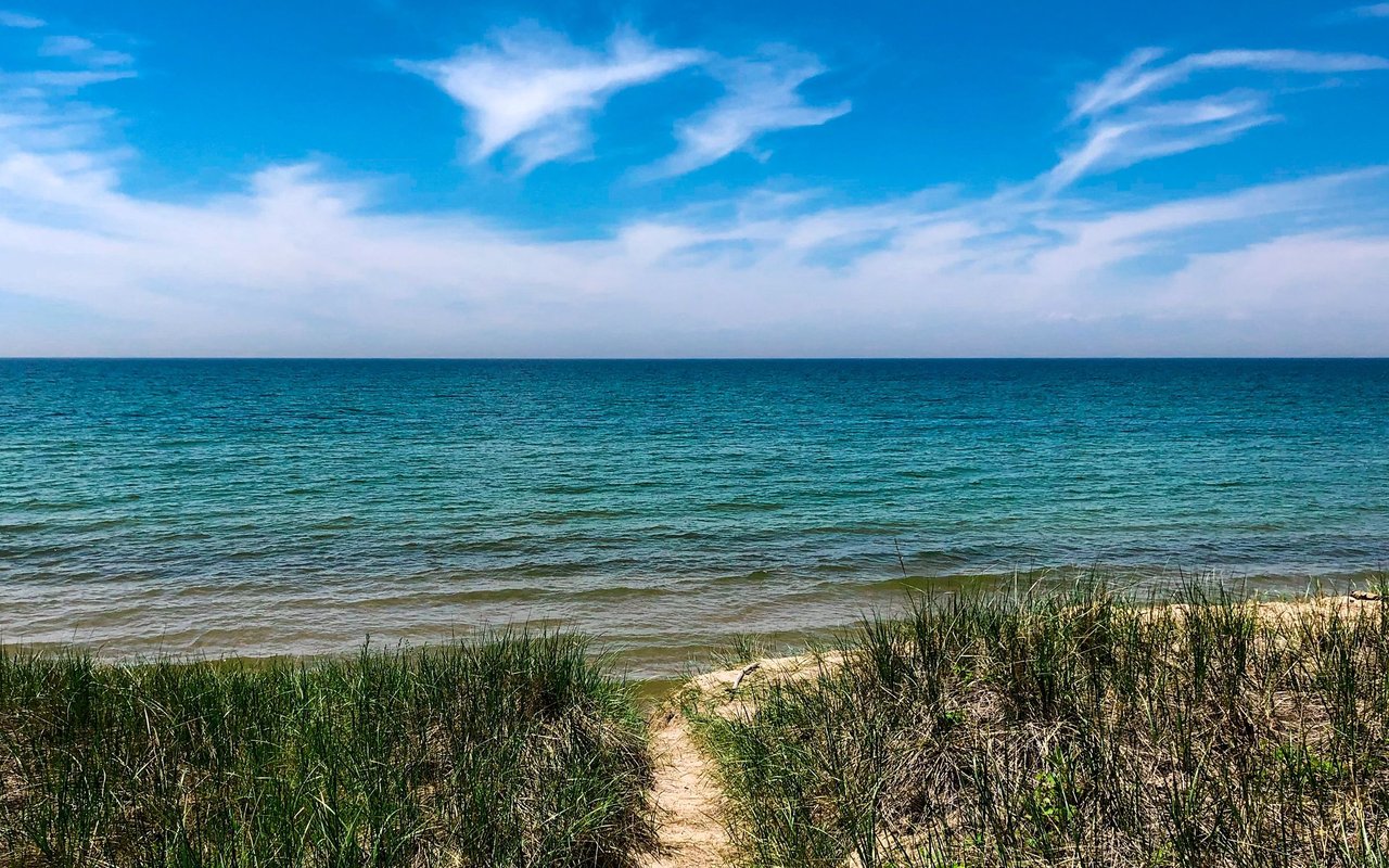 Sandy path through beach grass leading to a blue lake under a cloud-streaked sky.