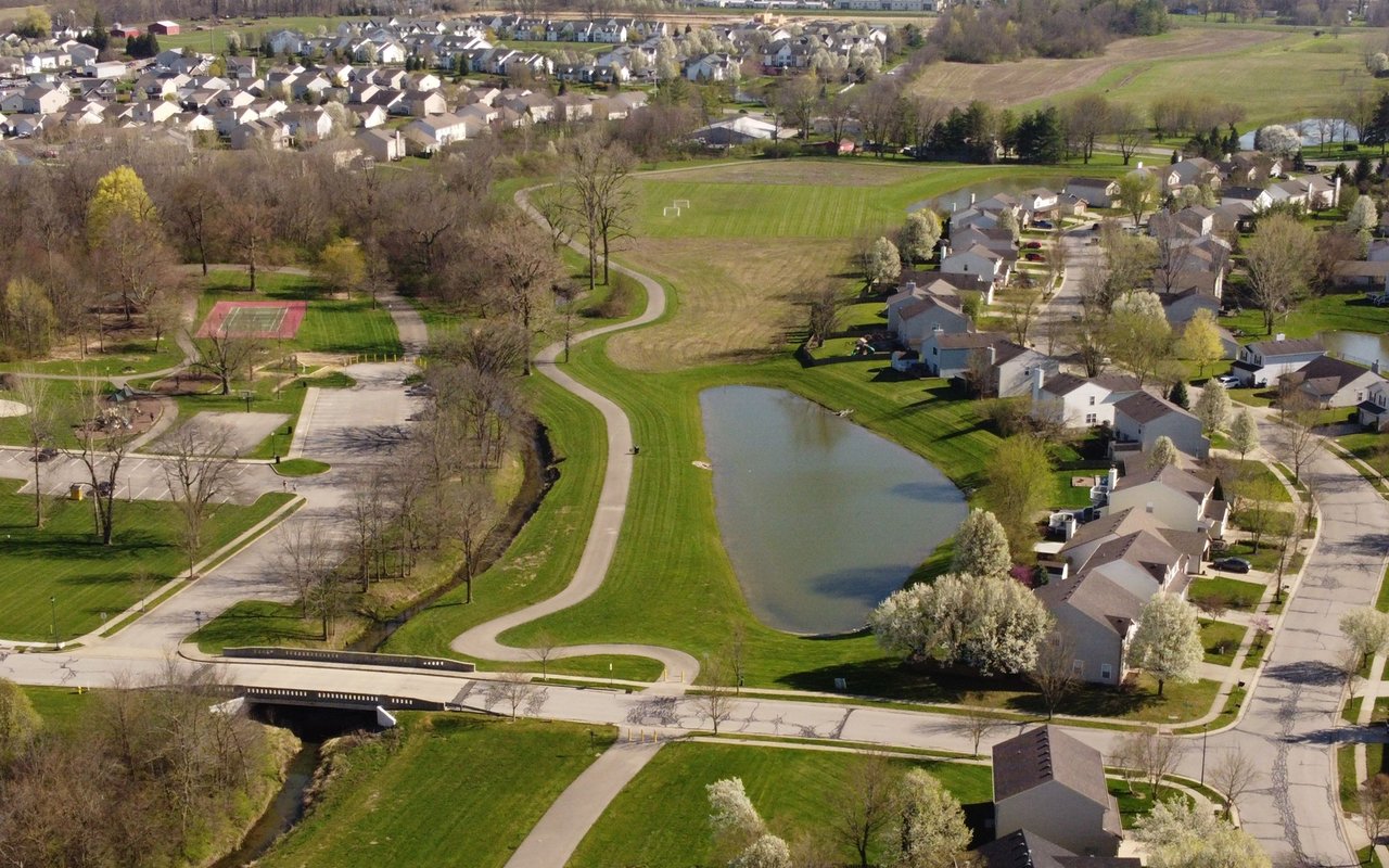 Aerial view of a neighborhood park with pond, walking paths, tennis court, and nearby homes.