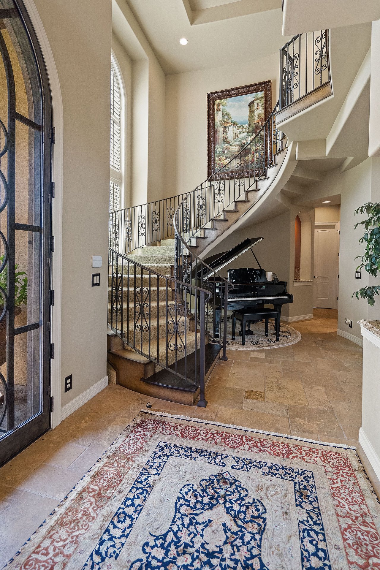 Foyer with a curved staircase and wrought iron railing, with a view to a piano beyond, at 25715 Cliff Circle, Lakecliff on Lake Travis, Spicewood, Texas.