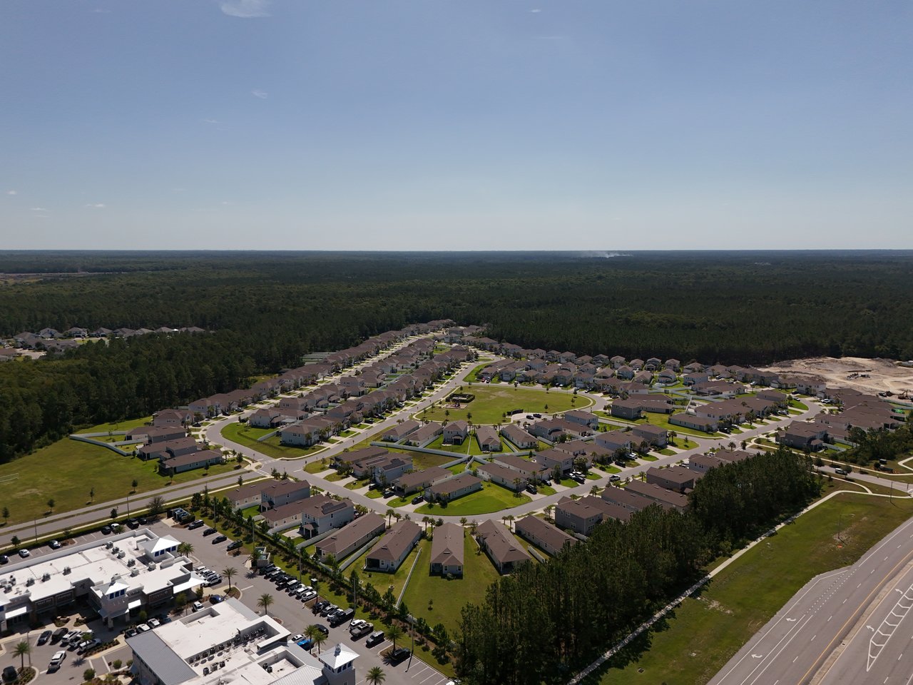 Aerial view of a neighborhood with rows of homes, wooded areas, and a retail complex beside a major road.