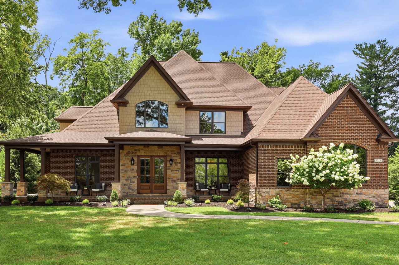 Two-story brick and stone home with a covered porch, steep roof, and flowering shrub.