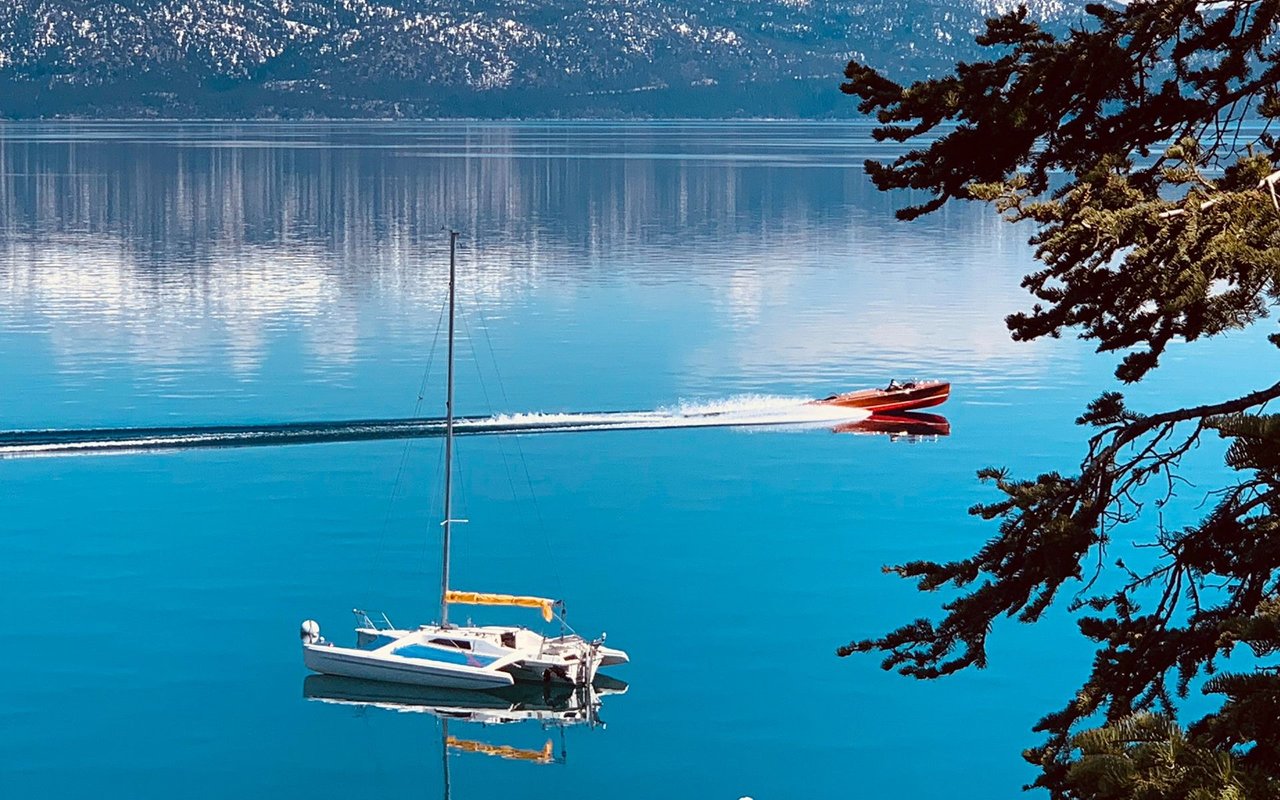 A red motorboat crosses Lake Tahoe past a moored sailboat, with mountains in the distance.