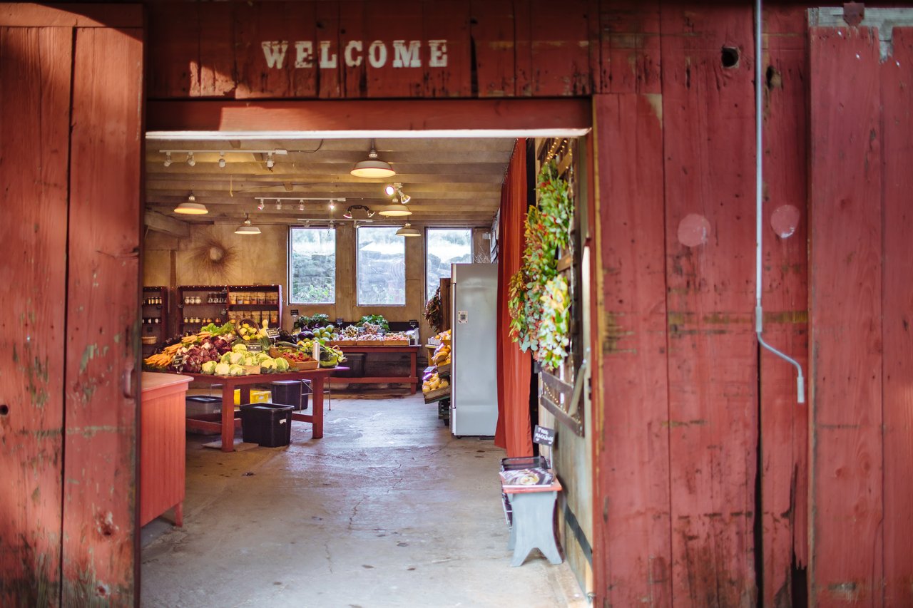 Small produce market viewed through a weathered red doorway marked “WELCOME.”