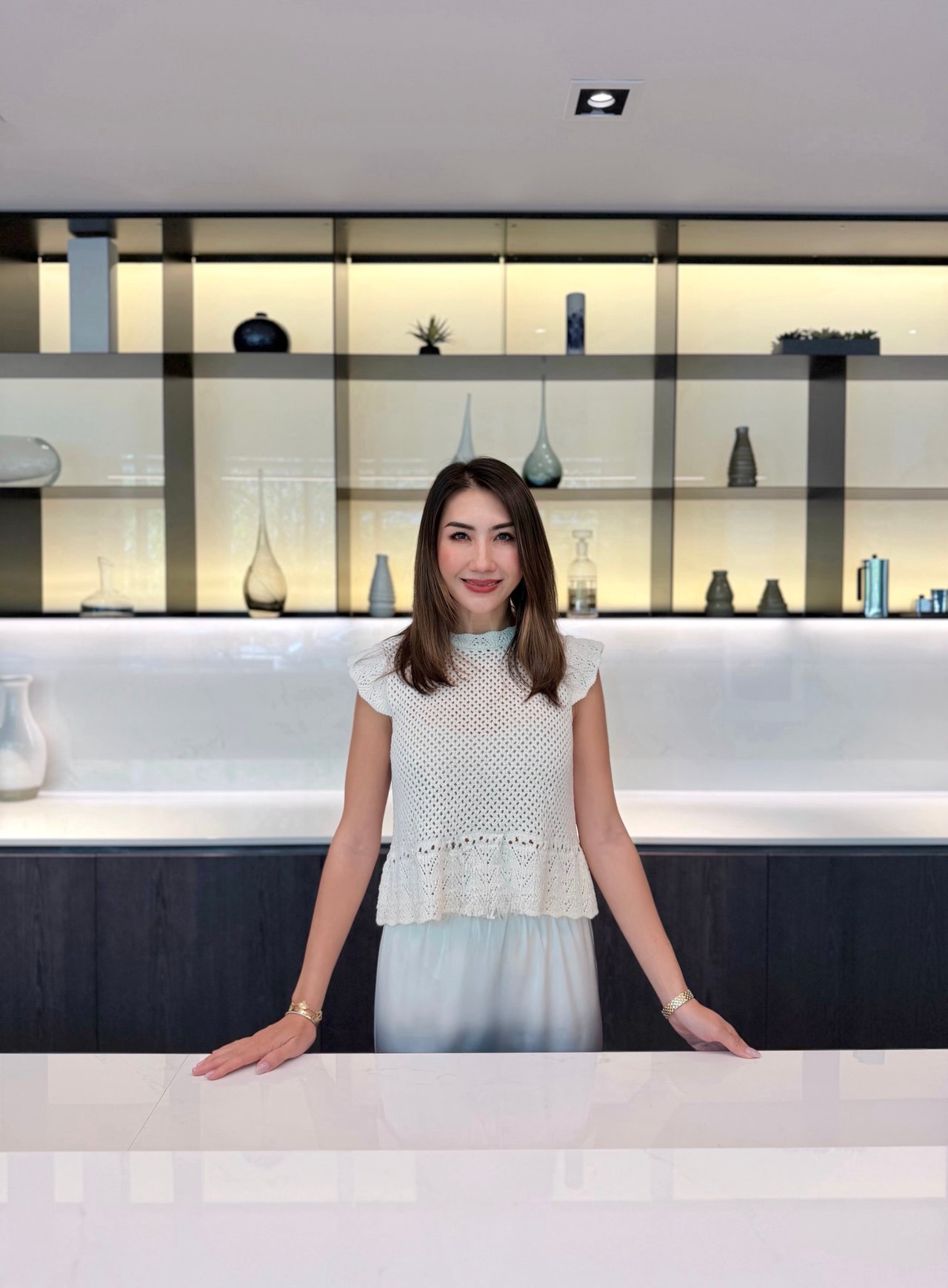 Leona Li poses behind a white marble counter in front of illuminated display shelves.