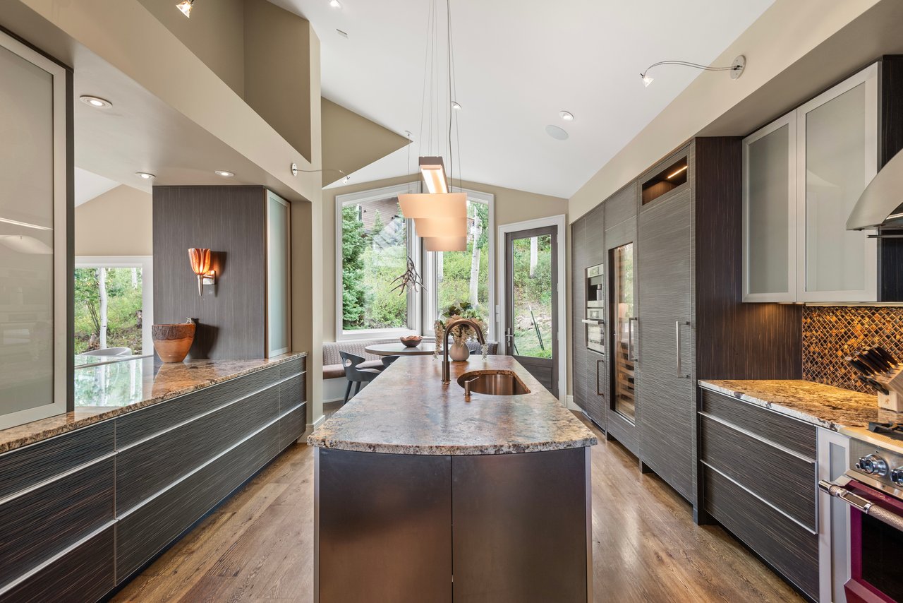 Modern kitchen with a large stone island, dark cabinetry, and windows overlooking trees.