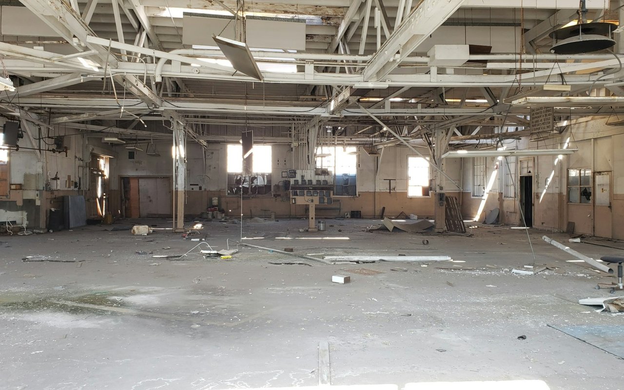 Vacant warehouse interior with exposed steel trusses and debris across the concrete floor.