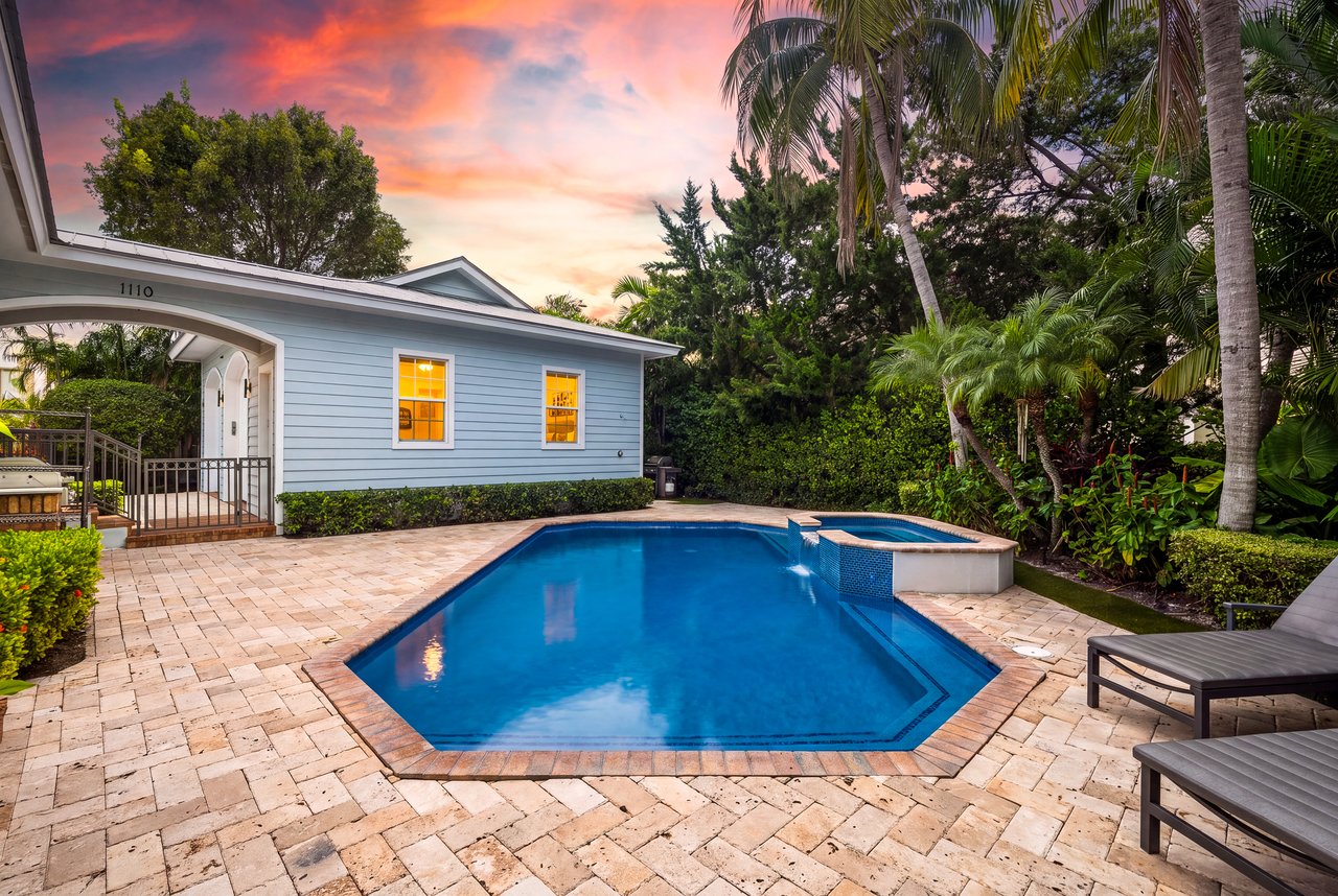 Backyard pool beside a light-blue house, framed by tropical palms at sunset.