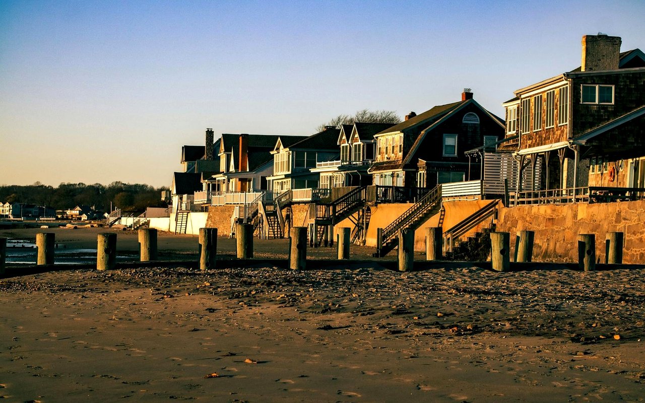 Coastal houses line a sandy beach above wooden pilings and seawalls.