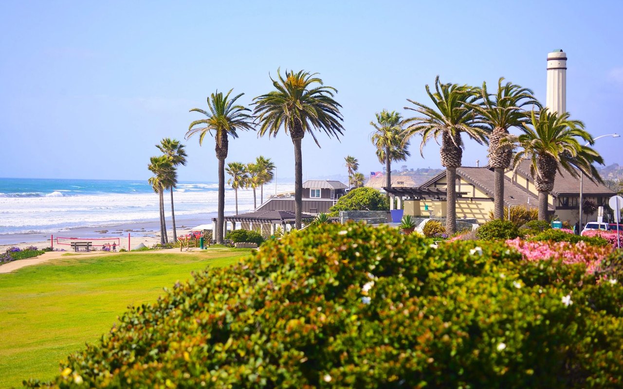 Del Mar coastline with ocean surf, palm trees, landscaped grounds, and beachside buildings.