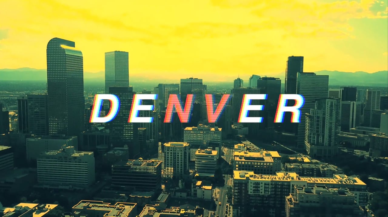 Aerial view of downtown Denver, with the skyline and distant mountains beneath a vivid yellow sky.