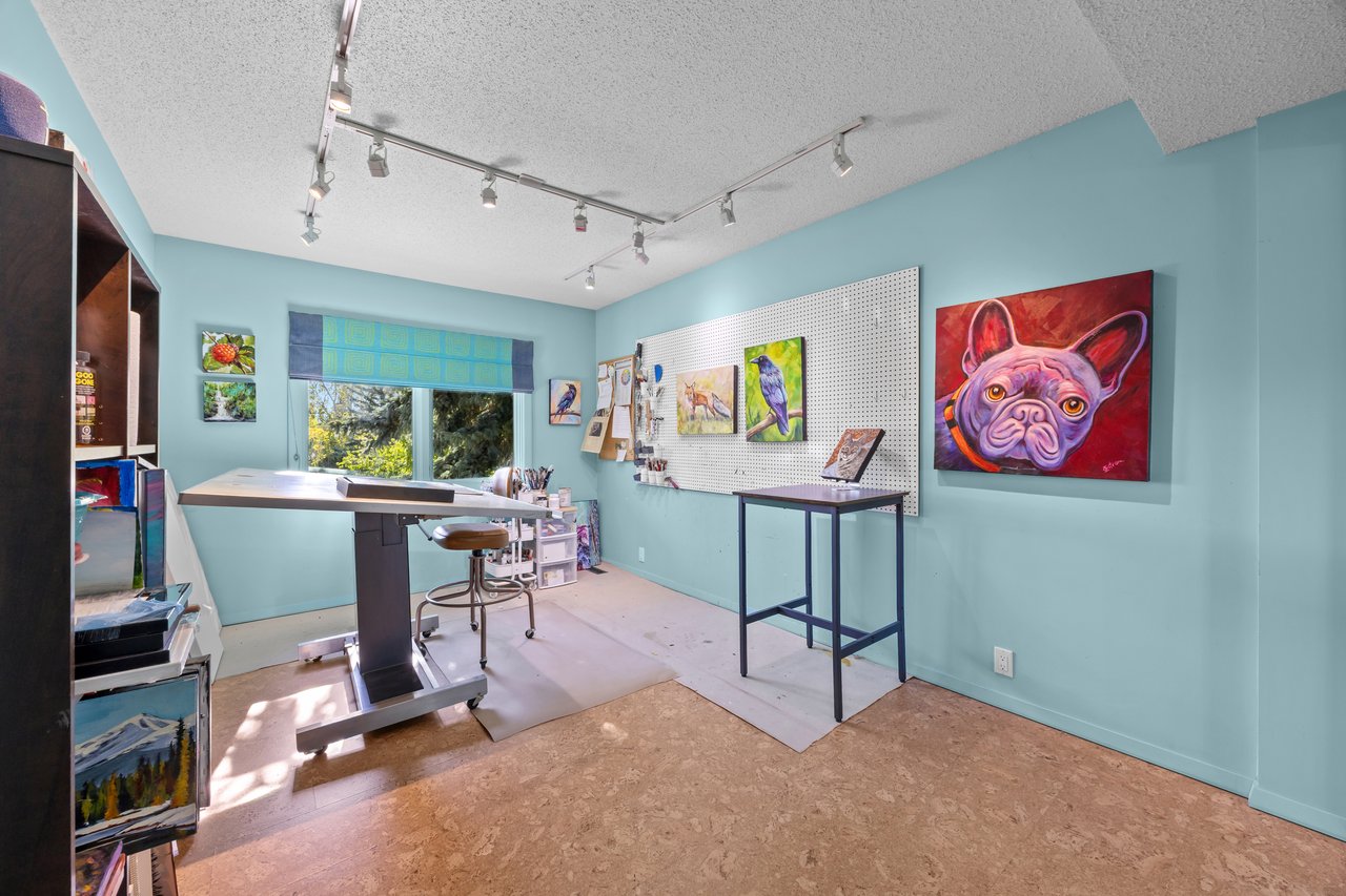 Art studio with a large worktable, colorful paintings, and a window overlooking trees.