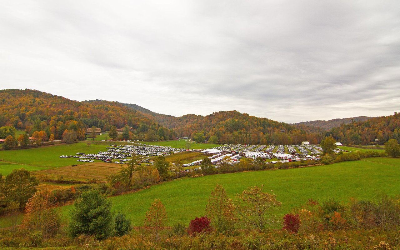 Autumn-colored mountain ridges overlook green fields and a large gathering of parked vehicles.