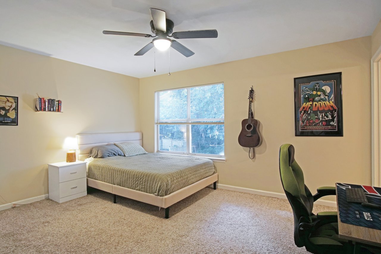 Bedroom at 529 Collinsford Road, Tallahassee, Florida, featuring beige walls, carpet flooring, and a ceiling fan with light fixture. The room includes a bed with green bedding, a nightstand with a lamp, a wall-mounted guitar, and a desk with a gaming chair. A large window provides natural light and views of the trees outside.