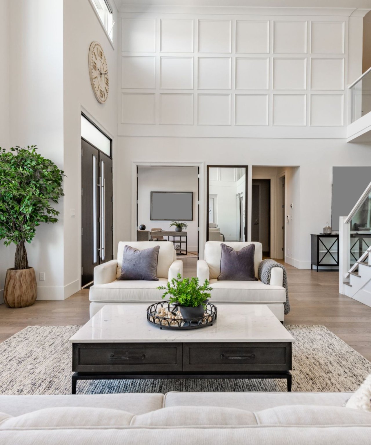 Double-height living room with white sofas, a paneled wall, marble-top coffee table, and glass-railed staircase.