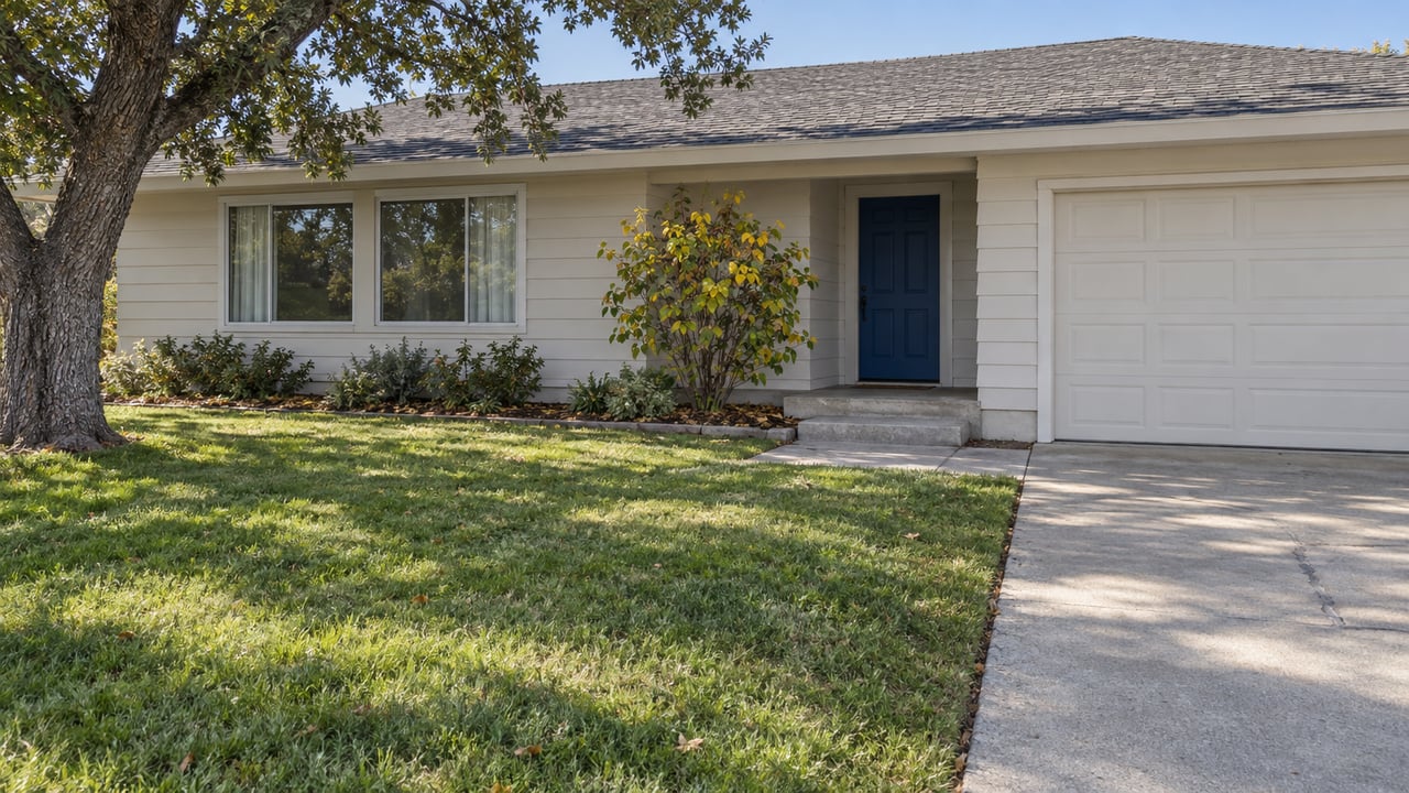 Single-story home with off-white siding, a blue front door, mature oak, deep lawn and concrete driveway in clear afternoon light.